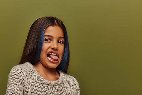 Portrait of stylish preteen girl with dyed hair wearing knitted sweater and sticking out tongue while looking at camera isolated on green, modern and hip preteen fashion concept