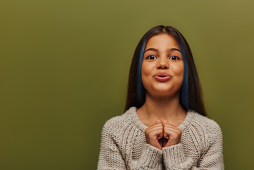 Excited stylish preteen girl with dyed hair in knitted sweater looking at camera and holding hands near chest while standing and posing isolated on green, stylish girl in cozy fall attire