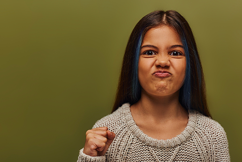 Portrait of angry and stylish preteen girl with dyed hair wearing knitted sweater while looking at camera and holding hand in fist isolated on green, stylish girl in cozy fall attire