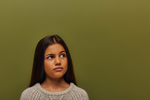 Portrait of thoughtful preadolescent brunette girl in stylish knitted sweater looking away while standing and posing isolated on green, stylish girl in cozy fall attire concept