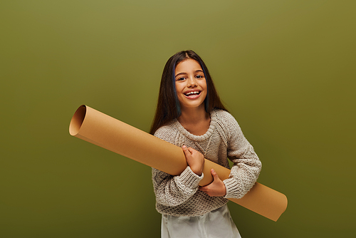 Cheerful and trendy brunette girl with dyed hair wearing modern knitted sweater holding rolled paper and looking at camera isolated on green, stylish girl in cozy fall attire concept