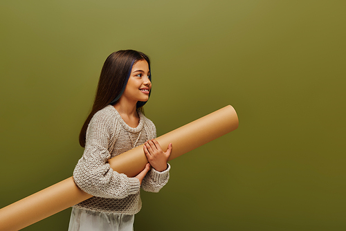 Smiling brunette preadolescent girl in trendy knitted sweater and outfit holding rolled paper and looking away isolated on green, stylish girl in cozy fall attire concept