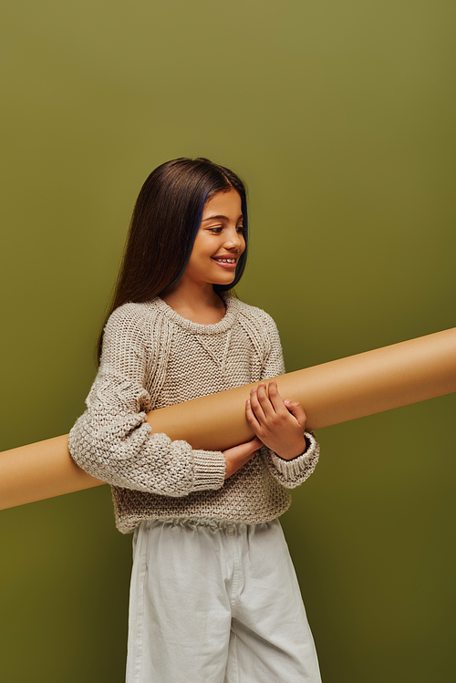 Cheerful brunette preadolescent girl in trendy autumn outfit and knitted sweater holding rolled paper while standing isolated on green, stylish girl in cozy fall attire concept