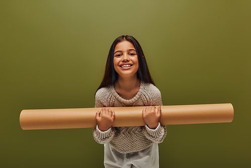 Positive brunette preteen girl in trendy autumn outfit and knitted sweater holding rolled paper and looking at camera while standing isolated on green, stylish girl in cozy fall attire