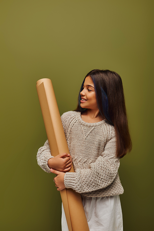 Joyful preadolescent with dyed hair wearing stylish and cozy autumn knitted sweater looking at rolled paper and standing isolated on green, girl radiating autumn vibes concept