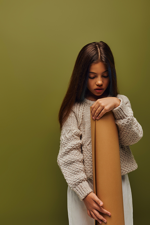 Stylish preadolescent child with dyed hair wearing cozy and modern knitted sweater while holding rolled paper while standing isolated on green, girl radiating autumn vibes concept