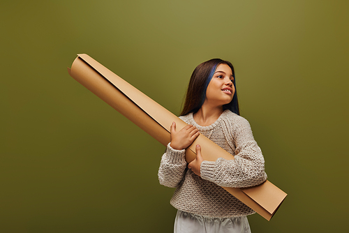 Smiling preadolescent girl with dyed hair wearing autumn outfit and knitted sweater while looking away and holding rolled paper isolated on green, girl radiating autumn vibes