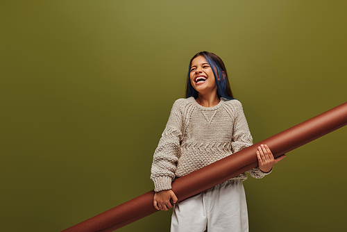 Excited and stylish preadolescent girl with dyed hair wearing cozy knitted sweater and holding rolled paper while standing isolated on green, girl radiating autumn vibes concept