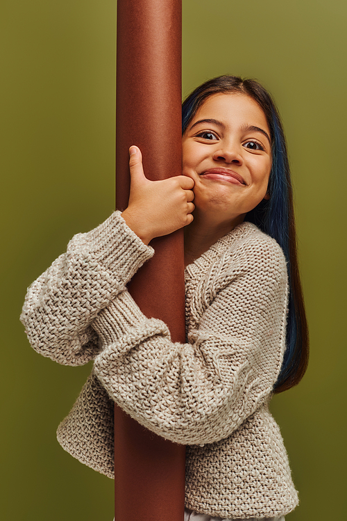 Portrait of excited and smiling trendy preteen girl in knitted sweater hugging rolled paper and looking at camera while standing isolated on green, girl radiating autumn vibes concept