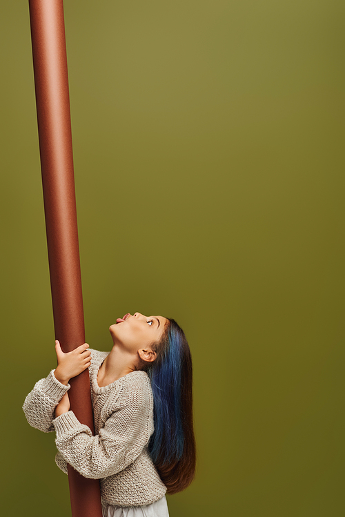 Side view of shocked and stylish preteen child with dyed hair wearing knitted sweater and looking up while holding rolled paper isolated on green, girl radiating autumn vibes