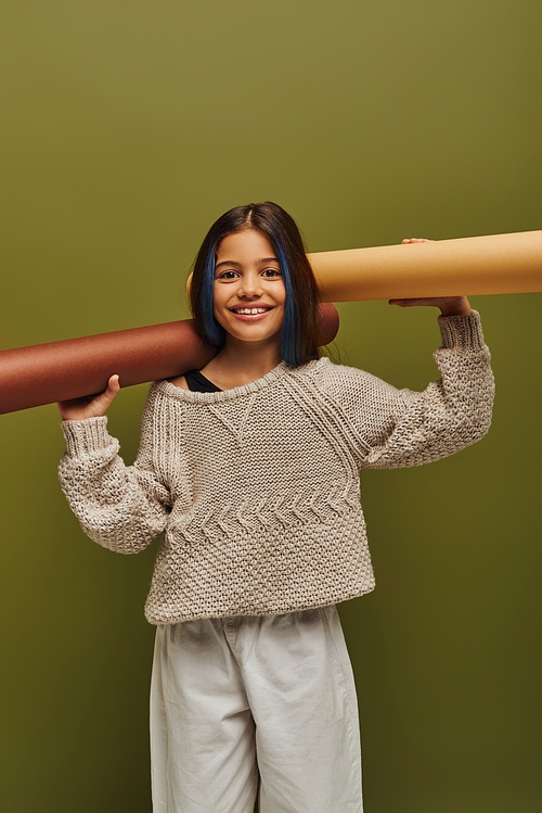 Cheerful preadolescent with dyed hair wearing autumn knitted sweater and looking at camera while holding rolled papers isolated on green, autumn fashion for preteens concept