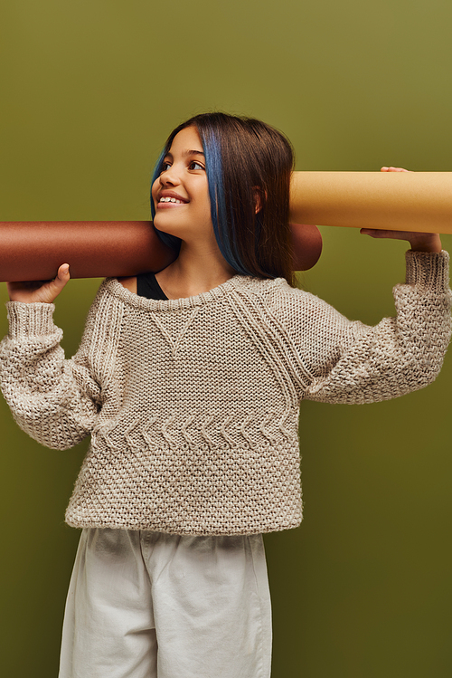 Positive preadolescent girl with dyed hair wearing trendy autumn outfit and holding rolled papers while looking away isolated on green, autumn fashion for preteens concept