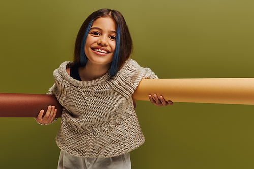 Positive preteen child with dyed hair wearing stylish knitted sweater and autumn outfit looking at camera and holding rolled papers isolated on green, autumn fashion for preteens concept