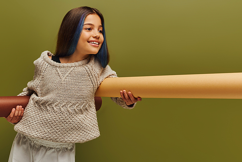 Smiling and trendy preadolescent girl with dyed hair wearing cozy knitted sweater and holding rolled papers and looking away isolated on green, autumn fashion for preteens concept