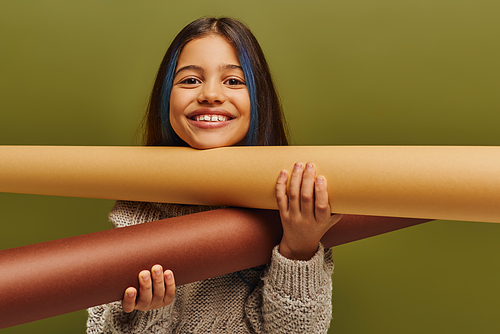 Portrait of positive preadolescent child with dyed hair wearing warm knitted sweater and holding rolled papers while standing isolated on green, autumn fashion for preteens concept