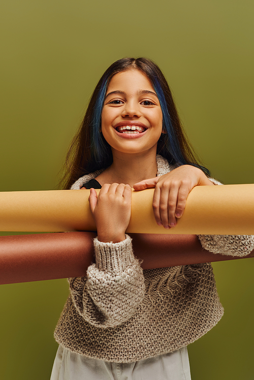 Cheerful preteen kid with dyed hair wearing knitted sweater and autumn outfit while holding rolled papers and standing isolated on green, autumn fashion for preteens concept