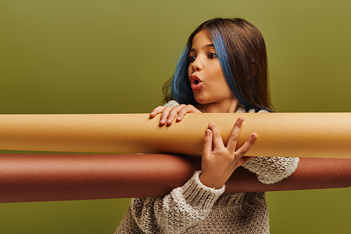 Shocked preadolescent girl with dyed hair wearing warm knitted sweater and holding rolled papers while standing and posing isolated on green, autumn fashion for preteens concept