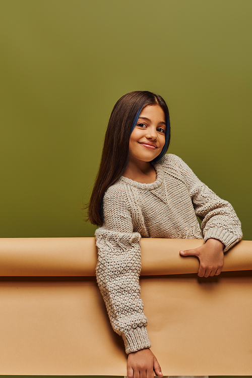 Joyful and stylish preadolescent girl with dyed hair wearing warm knitted sweater and looking at camera while holding rolled paper isolated on green, autumn fashion for preteens concept