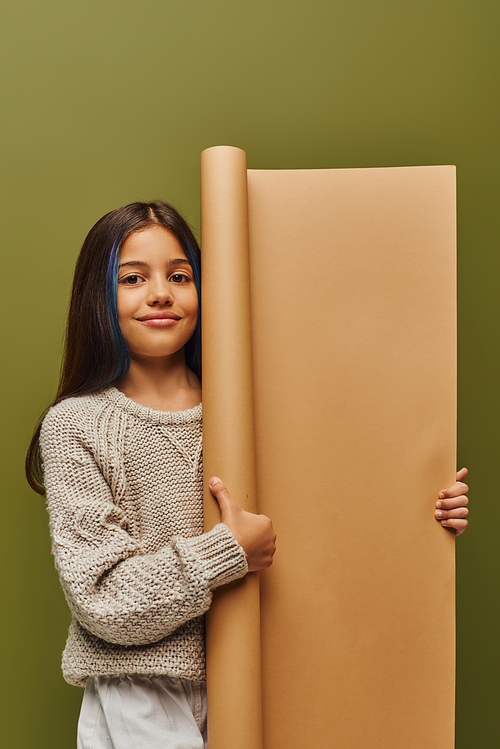 Smiling and trendy preadolescent girl with dyed hair wearing knitted sweater and autumn outfit and holding rolled paper isolated on green, autumn fashion for preteens concept