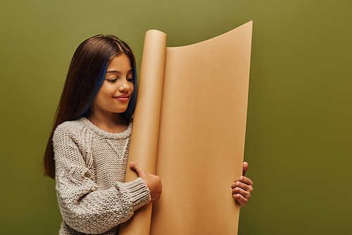 Smiling preteen girl with dyed hair and trendy cozy knitted sweater looking at rolled paper while standing and posing isolated on green, autumn fashion for preteens concept