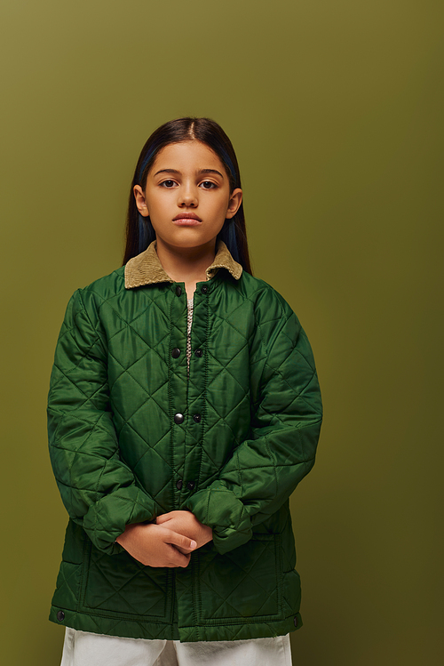 Serious and trendy preadolescent girl with dyed hair looking at camera while posing in autumn jacket isolated on green, modern fall fashion for preteens concept