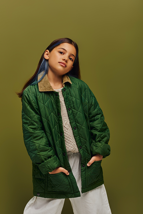 Confident and stylish brunette preteen girl with dyed hair posing in autumn outfit and knitted sweater and looking at camera isolated on green, modern fall fashion for preteens concept