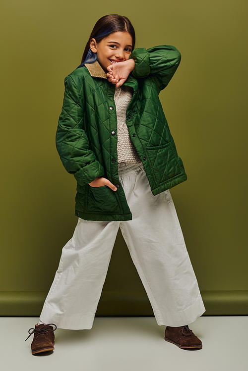 Full length of positive preadolescent girl with dyed hair looking at camera while posing in autumn outfit and knitted sweater on green background, modern fall fashion for preteens concept