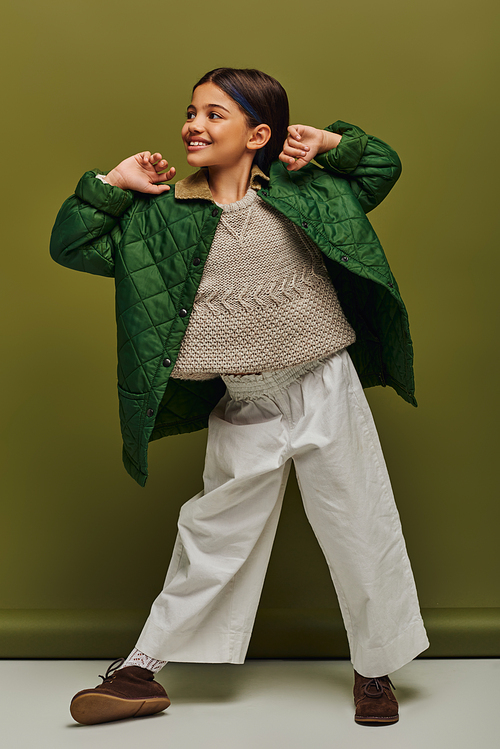 Full length of smiling and trendy preteen kid in autumn outfit and knitted sweater looking away while posing and standing on green background, modern child fashion for preteens concept