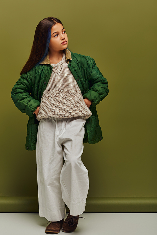 Full length of trendy preadolescent girl with dyed hair and warm knitted sweater holding hands on hips and posing on green background, modern fashion for preteens concept