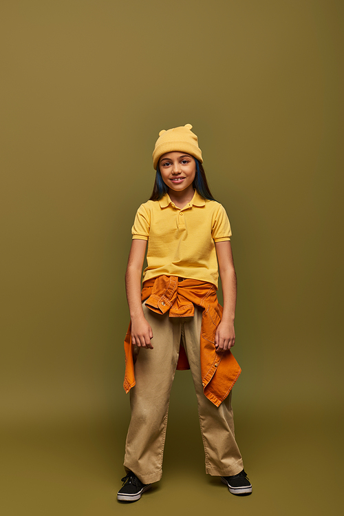 Full length of smiling and stylish preadolescent girl with dyed hair wearing urban outfit and yellow hat while standing on khaki background, stylish girl in modern outfit concept