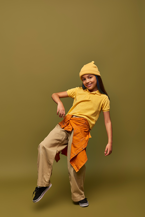 Full length of trendy and cheerful preadolescent girl in urban outfit and yellow hat posing while looking at camera on khaki background, stylish girl in modern outfit concept