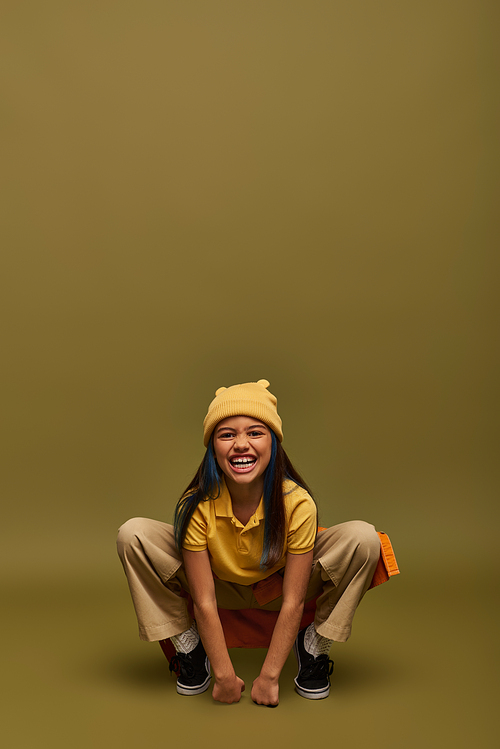 Excited and trendy preteen girl with dyed hair posing in yellow hat and urban outfit while standing and looking at camera on khaki background, stylish girl in modern outfit concept