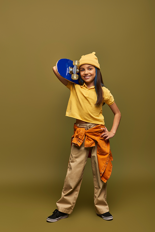 Full length of cheerful and stylish preteen girl with dyed hair wearing urban outfit and yellow hat and holding skateboard on khaki background, stylish girl in modern outfit concept