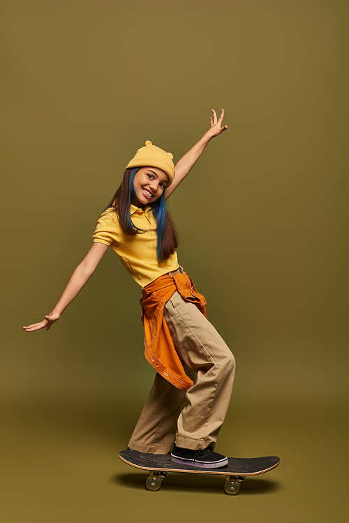 Positive and stylish preadolescent girl with dyed hair wearing yellow hat and urban outfit while looking at camera near skateboard on khaki background, stylish girl in modern outfit concept