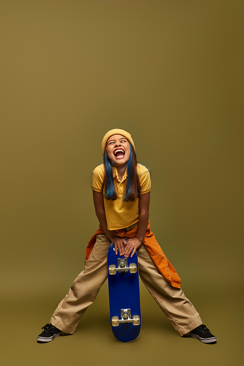 Full length of excited preteen child with colored hair wearing urban outfit and hat while grimacing and holding skateboard on khaki background, stylish girl in modern outfit concept