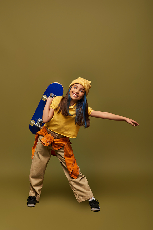 Full length of positive preteen girl with dyed hair wearing yellow hat and urban outfit while holding skateboard and standing on khaki background, stylish girl in modern outfit concept