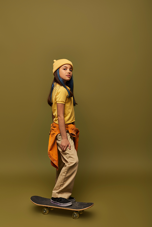 Confident preteen girl with dyed hair wearing hat and urban outfit while looking at camera and standing on skateboard on khaki background, girl with cool street style look