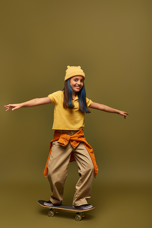 Joyful preadolescent girl with dyed hair wearing stylish urban outfit and hat while standing on skateboard and looking at camera on khaki background, girl with cool street style look