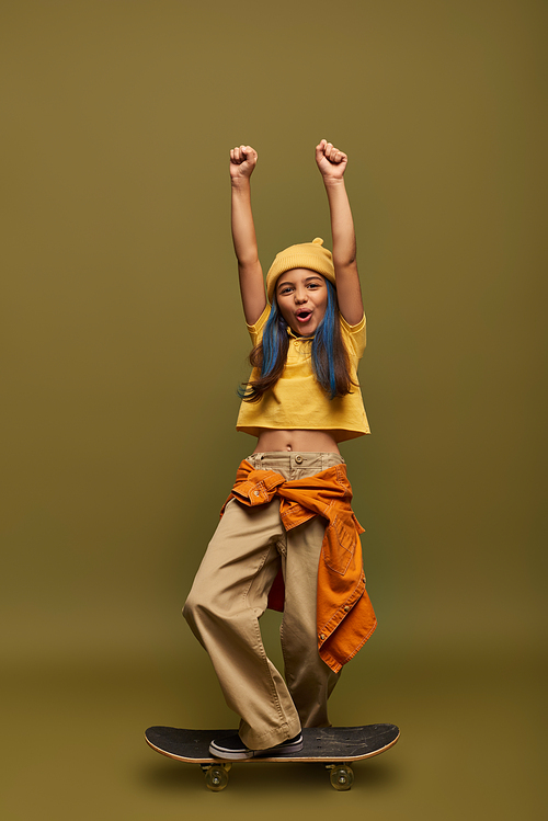 Full length of excited and stylish preteen girl with dyed hair showing yes gesture and looking at camera while standing on skateboard on khaki background, girl with cool street style look