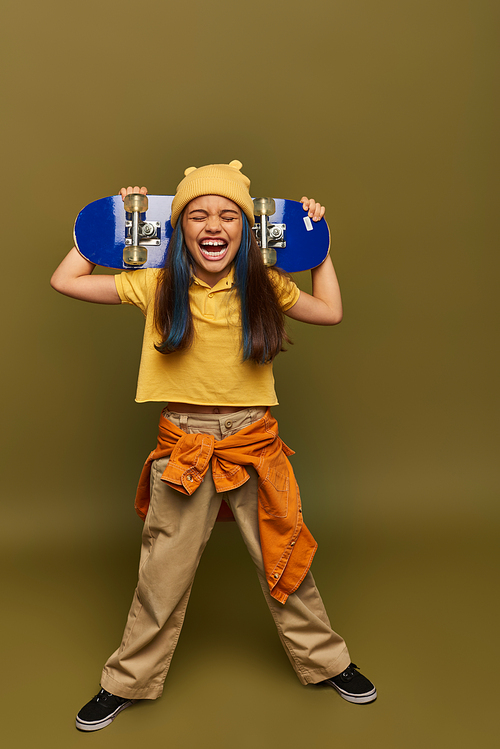 Full length of excited preteen kid with dyed hair wearing hat and stylish urban outfit laughing and holding skateboard on khaki background, girl with cool street style look