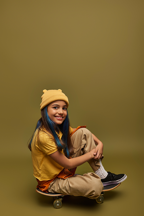 Smiling preteen and stylish child with dyed hair wearing urban outfit and hat looking at camera while sitting on skateboard on khaki background, girl with cool street style look