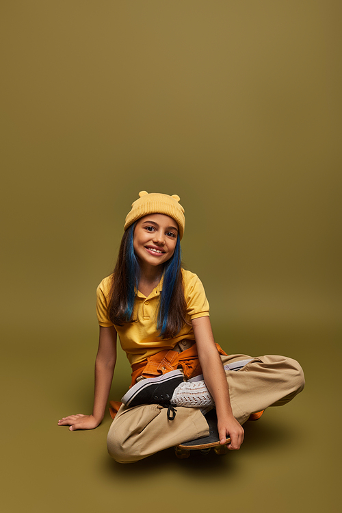 Positive and fashionable preteen girl with dyed hair wearing urban clothes and yellow hat while looking at camera and sitting on skateboard on khaki background, girl with cool street style look