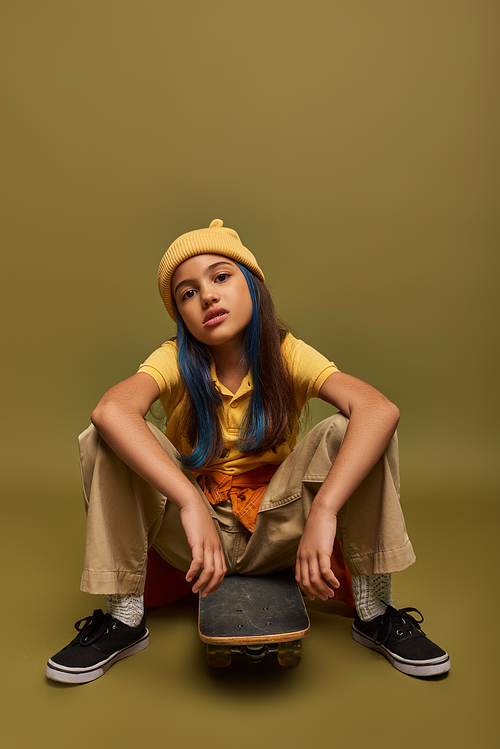 Full length of confident and serious preteen girl with dyed hair wearing urban outfit and hat and sitting on skateboard and looking at camera on khaki background, girl with cool street style look