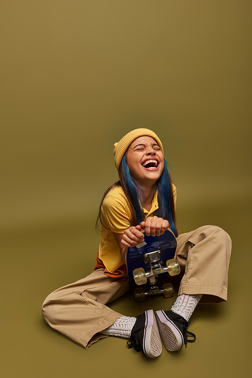 Laughing and stylish preadolescent girl with colored hair wearing urban outfit and yellow hat holding skateboard and sitting on khaki background, girl with cool street style look