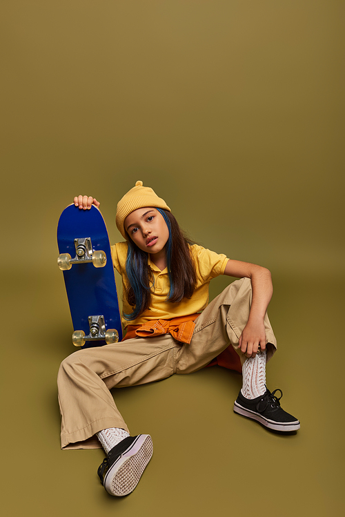 Full length of confident and fashionable preadolescent child with colored hair wearing urban clothes and hat while holding skateboard on khaki background, girl with cool street style look