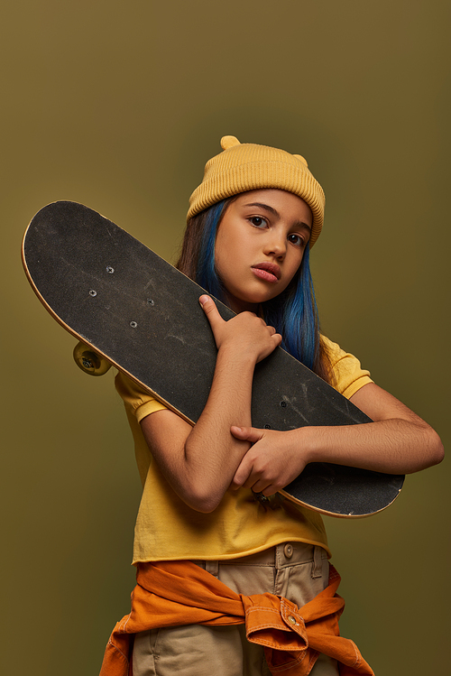 Portrait of stylish preadolescent girl in yellow hat and urban outfit looking at camera while holding skateboard and posing isolated on khaki, girl in urban streetwear concept