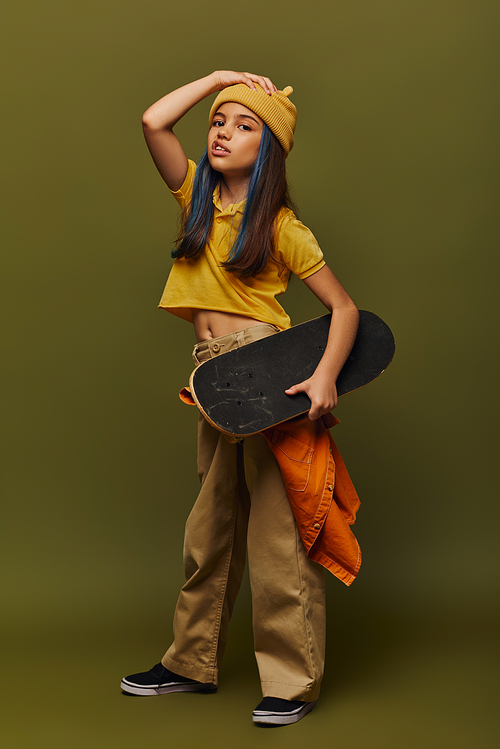 Full length of trendy preteen girl with dyed hair wearing urban outfit and holding skateboard while grimacing and looking at camera on khaki background, girl in urban streetwear concept