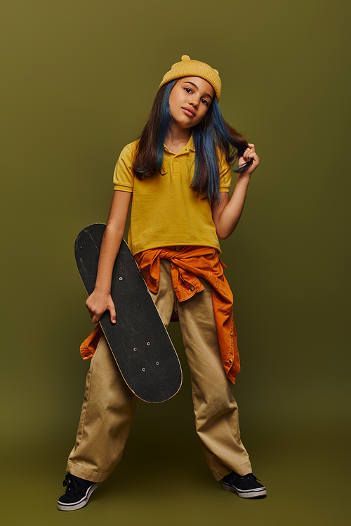 Full length of confident and fashionable preteen girl with dyed hair wearing hat and urban outfit and holding skateboard on khaki background, girl in urban streetwear concept