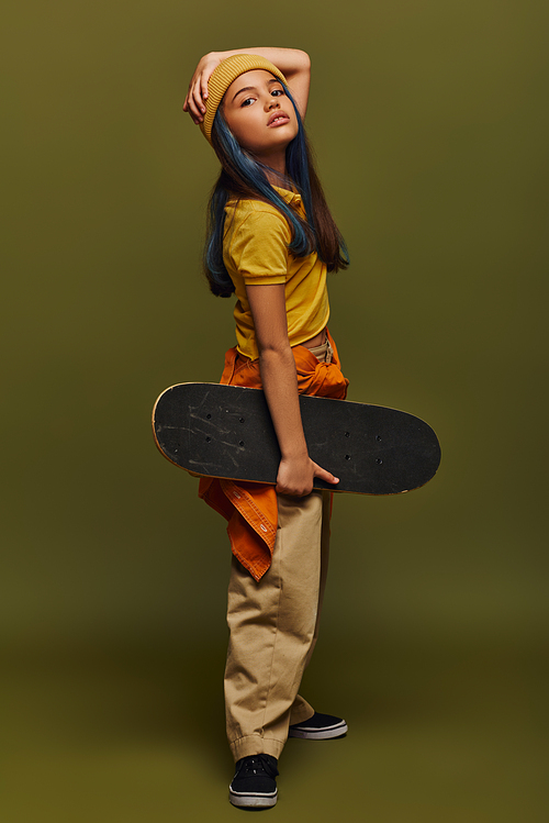 Full length of trendy and confident preadolescent kid with colored hair wearing hat and urban outfit and holding skateboard while standing on khaki background, girl in urban streetwear concept