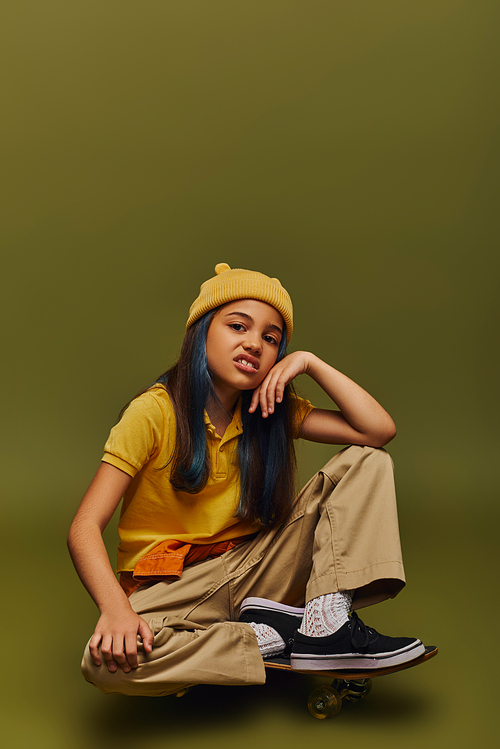 Disgusted and fashionable preteen girl with dyed hair wearing yellow hat and urban outfit while looking at camera and sitting on skateboard on khaki background, girl in urban streetwear concept